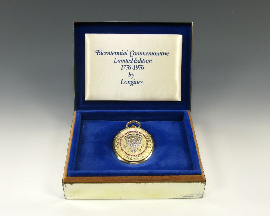 Silver pocket watch has the Seal of the President depicted on the front set with colored stones. The watch face features a silver background with gold details. The pocket watch sits inside a silver box lined with blue velvet. Gift of Betty Ford.